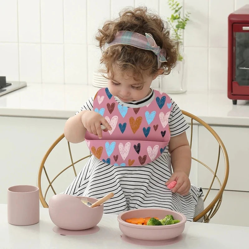 Petite fille assise à table avec un bavoir bébé silicone rose décoré de cœurs colorés