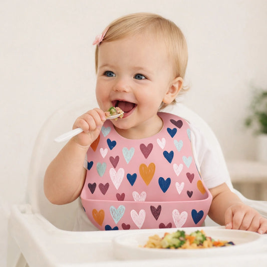 bébé fille souriante mange avec cuillère portant bavoir silicone rose coeurs imperméable avec récupérateur
