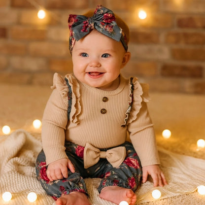Bébé fille en tenue beige foncé avec pantalon fleuri et grand nœud, assise parmi des lumières de Noël chaleureuses