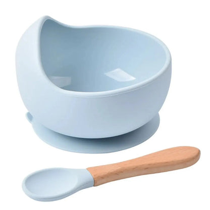 Bol en silicone bleu pastel pour bébé avec rebord surélevé, base à ventouse et cuillère assortie à manche en bois sur fond blanc.