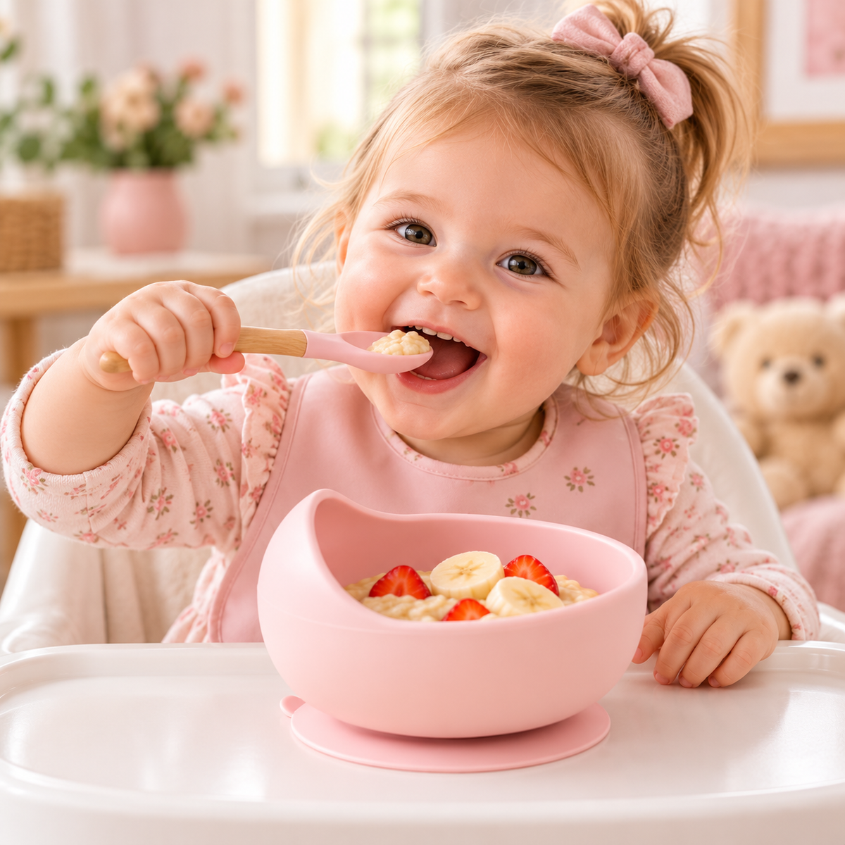 Petite fille souriante mangeant avec une cuillère rose dans un bol en silicone rose à ventouse, garni de fruits et de céréales.