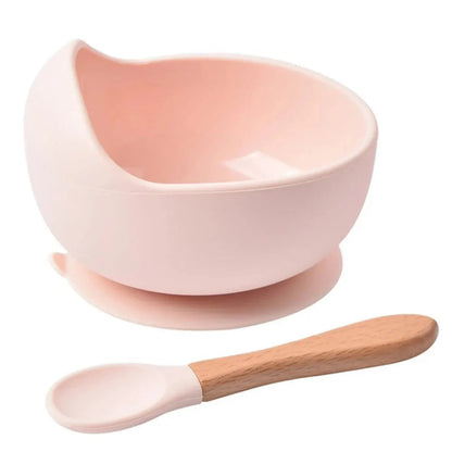 Bol en silicone rose pastel pour bébé avec rebord haut, ventouse et cuillère rose à manche en bois, présenté sur fond blanc.
 Copier