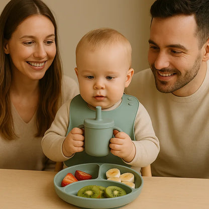 Bébé avec bavoir en silicone tenant un gobelet, assiette de fruits et parents souriants