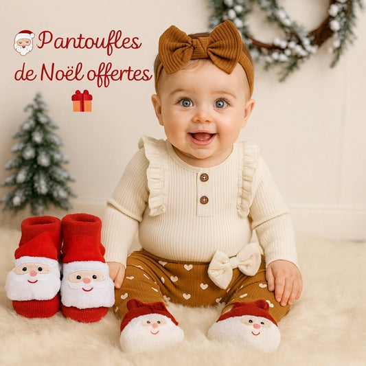 Bébé fille souriante en tenue beige et marron avec bandeau et pantoufles Père Noël, décor de Noël chaleureux avec sapins – pantoufles de Noël offertes par Cigogne & Bébé.